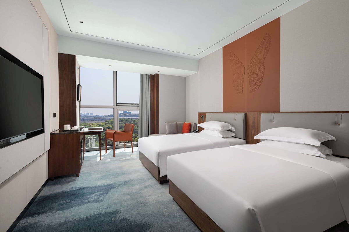 A hotel room with two beds, a desk, a TV, and a view of the city.