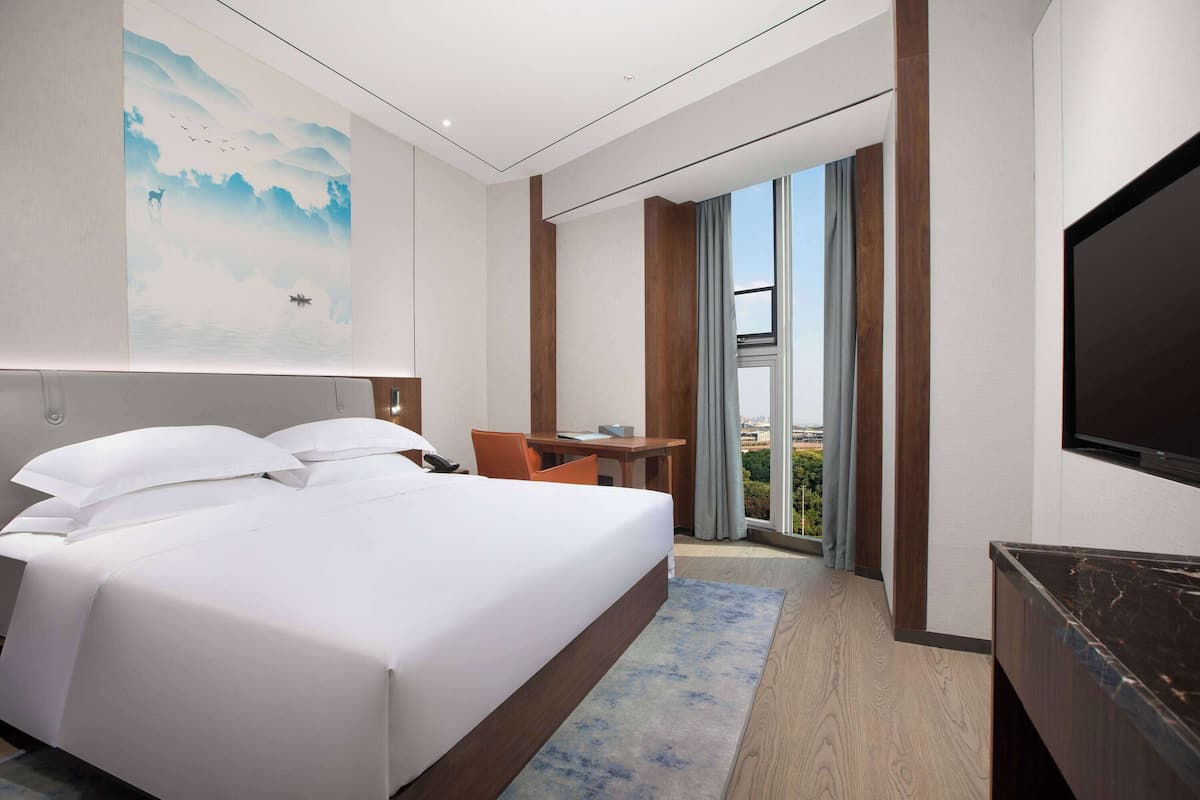 A modern hotel room with a large bed, a desk, a chair, a TV, and a large window with a city view.