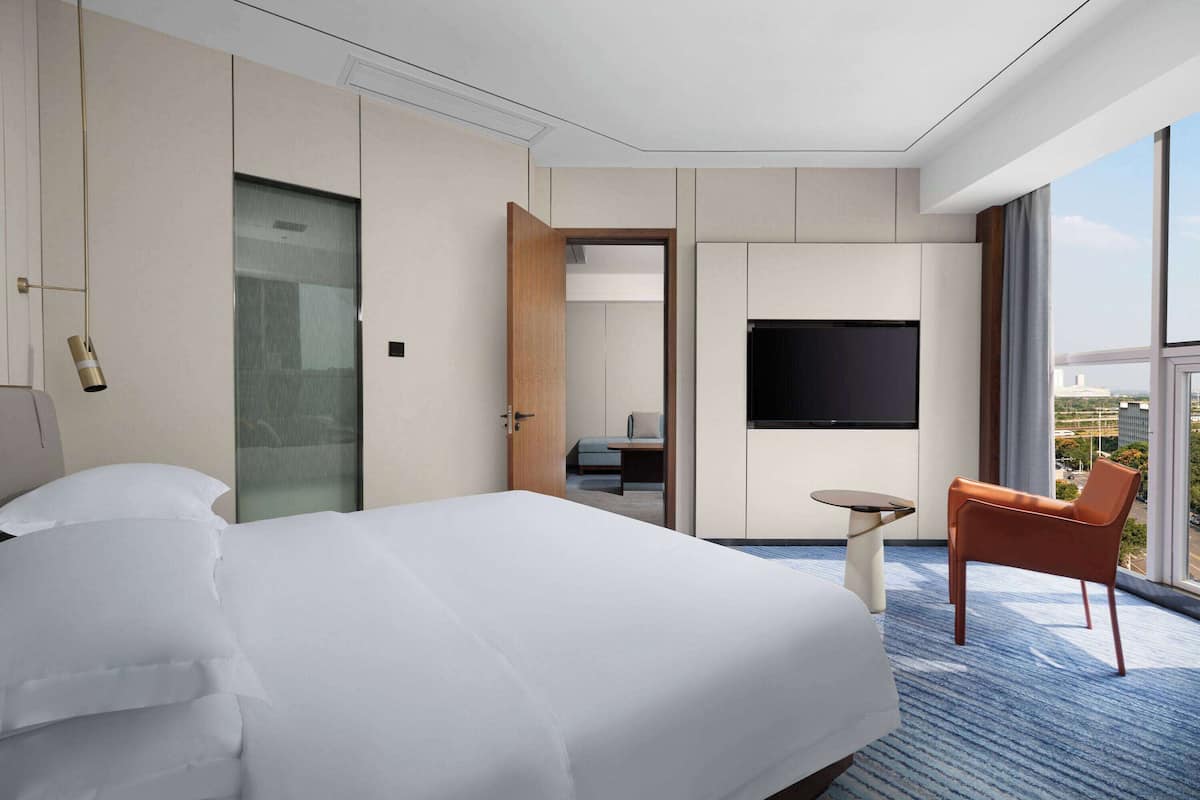 A modern hotel room with a large bed, a flat-screen TV, a small table, and a window with a city view.
