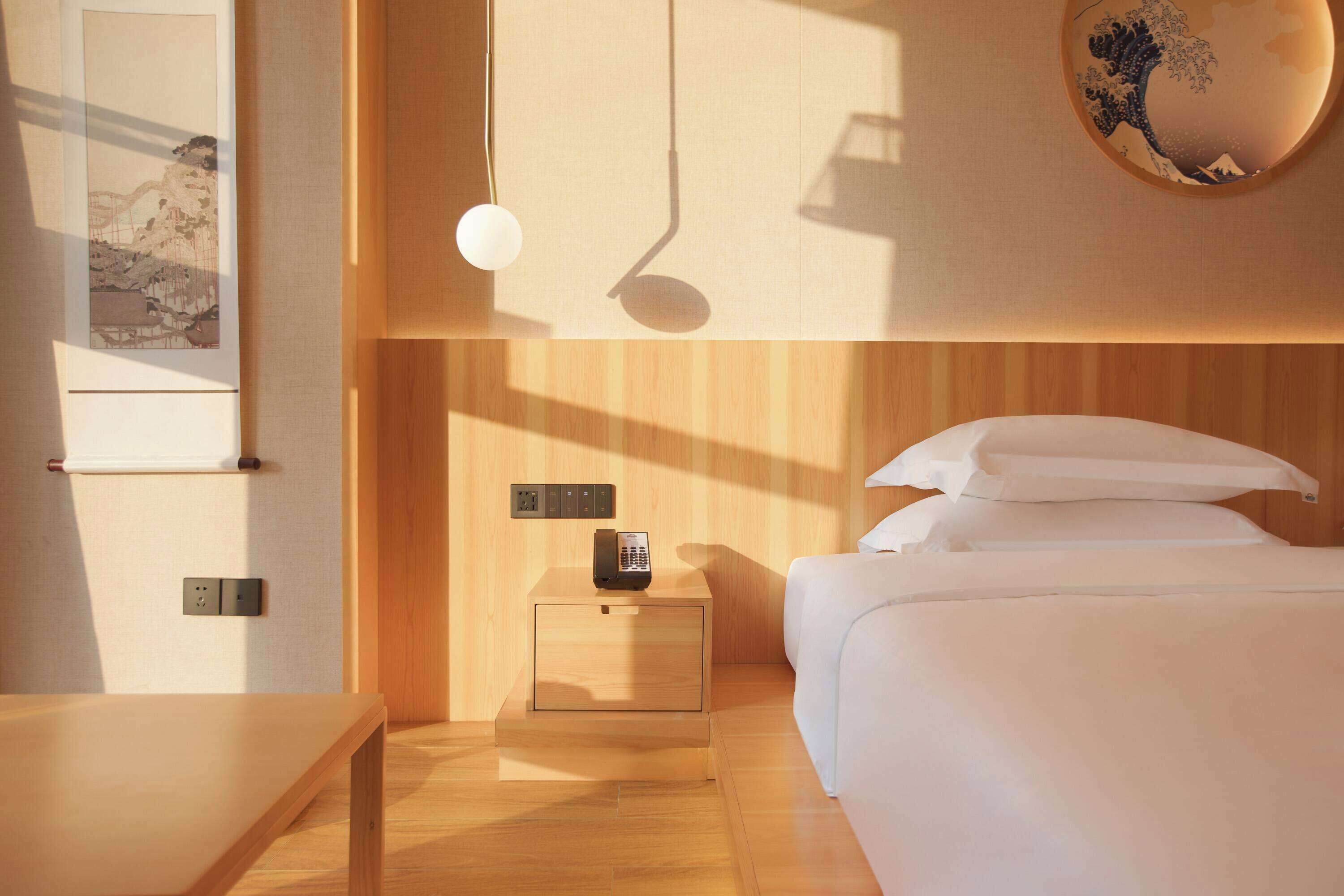 Une chambre d’hôtel avec un plancher en bois, un lit avec du linge de maison blanc, une table de chevet avec un téléphone et un luminaire fixé au mur.