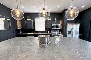 Une cuisine moderne dotée d’un grand îlot central, d’appareils électroménagers en acier inoxydable et de luminaires suspendus.