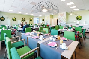 A dining area with tables set for breakfast, green upholstered chairs, and a ceiling fan. - Hotel Am Zoo (Neunkirchen)