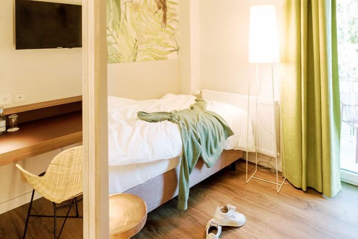 A hotel room with a bed, a desk with a chair, a TV mounted on the wall, and a window with green curtains.