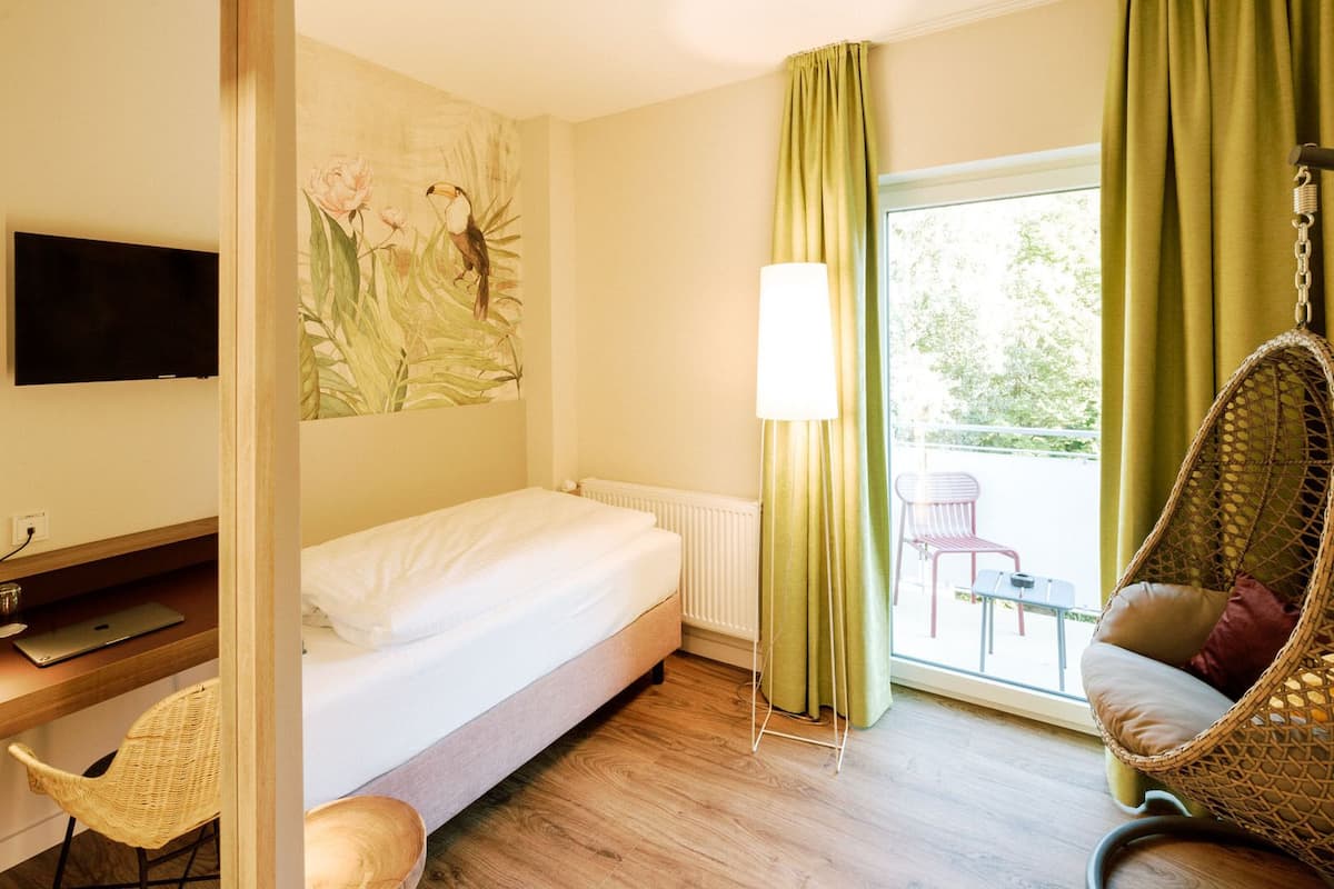 A hotel room with a bed, a desk with a laptop, a chair, a hanging chair, and a window with curtains.