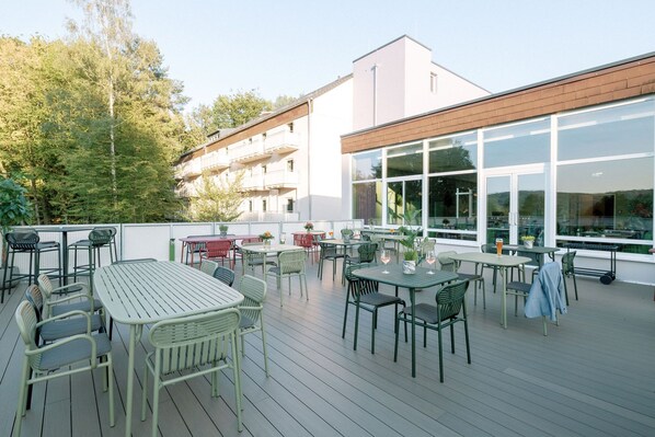 An outdoor patio with multiple tables and chairs, a wooden deck, and a building with large windows in the background. - Hotel Am Zoo (Neunkirchen)