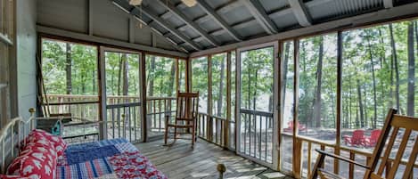 Una habitación acogedora con una cama, ventilador de techo y amplias ventanas con vistas al bosque.