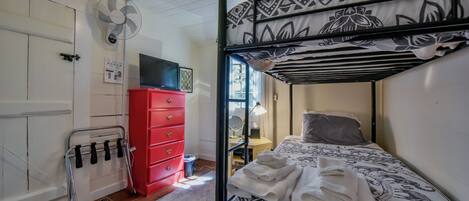 Una cama litera con colcha estampada, un aparador rojo, un televisor y un ventilador fijado a la pared en una habitación con paredes blancas y suelo de madera.
