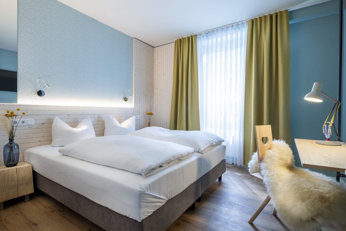 Quarto de hotel moderno com cama grande, piso de madeira, escrivaninha com luminária e uma cadeira.