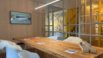 Un salón de conferencias con una mesa de madera, sillas blancas, una pantalla montada y un panel de vidrio con un mural colorido.