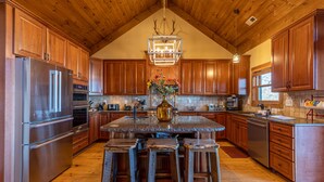 A spacious kitchen with wooden cabinets, a large island with stools, stainless steel appliances, and a chandelier. - Buckhorn Lodge - Spacious Luxury Home Near Sugar Mountain Resort, Pool Table, Hot Tub, Views (Sugar Mountain)