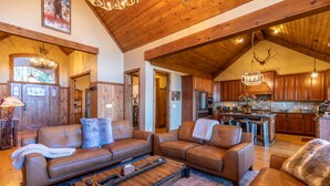 A spacious living room with a large brown leather sofa, a wooden coffee table, and a kitchen area with wooden cabinets and a hanging light fixture. - Buckhorn Lodge - Spacious Luxury Home Near Sugar Mountain Resort, Pool Table, Hot Tub, Views (Sugar Mountain)