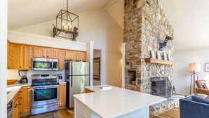 Una cocina moderna con armarios de madera, electrodomésticos de acero inoxidable, una chimenea de piedra y una zona de estar acogedora con un sofá azul y una mesa de madera.