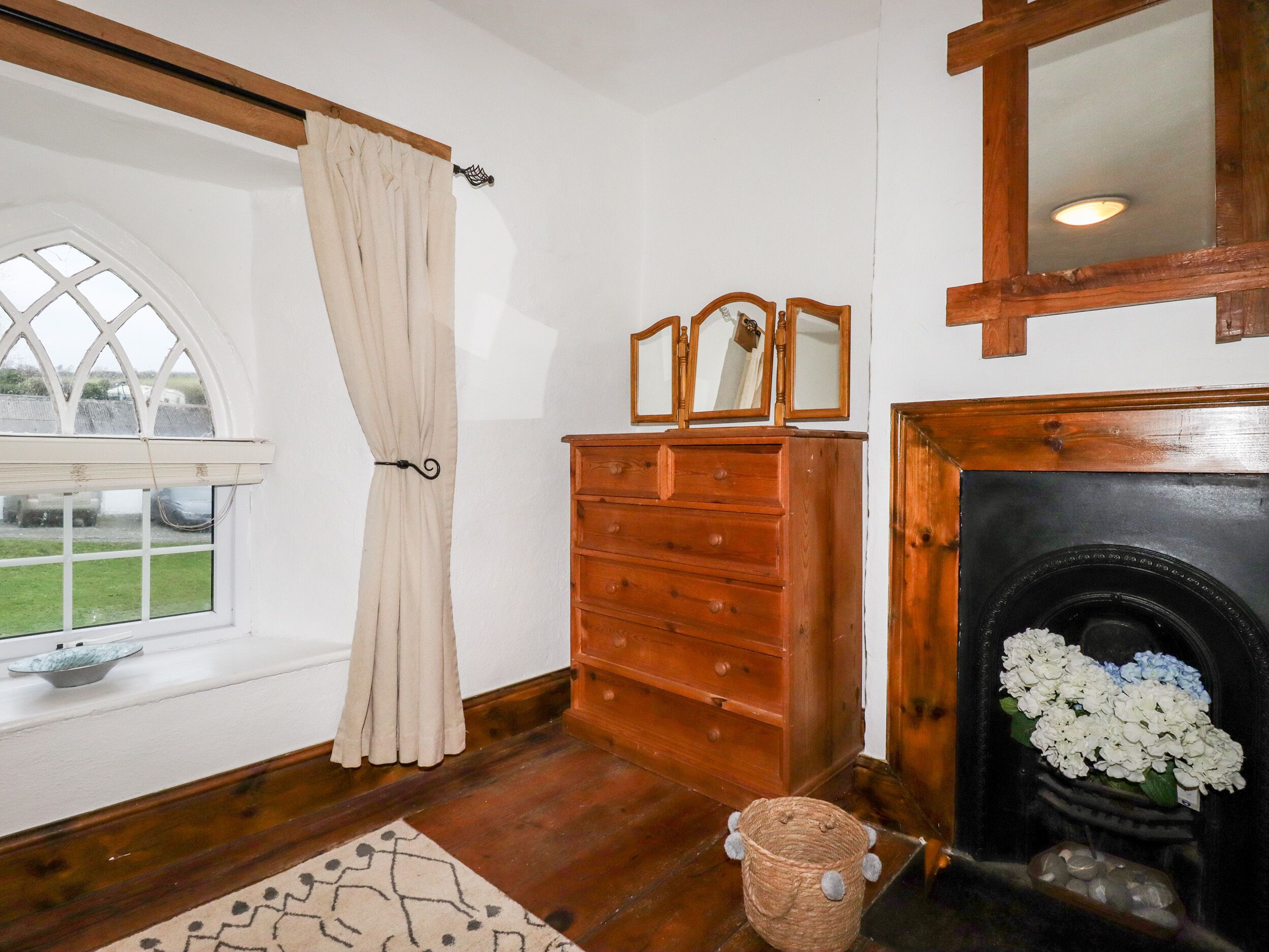 A room with a wooden dresser, a mirror, a fireplace with white flowers, and a window with a curtain.