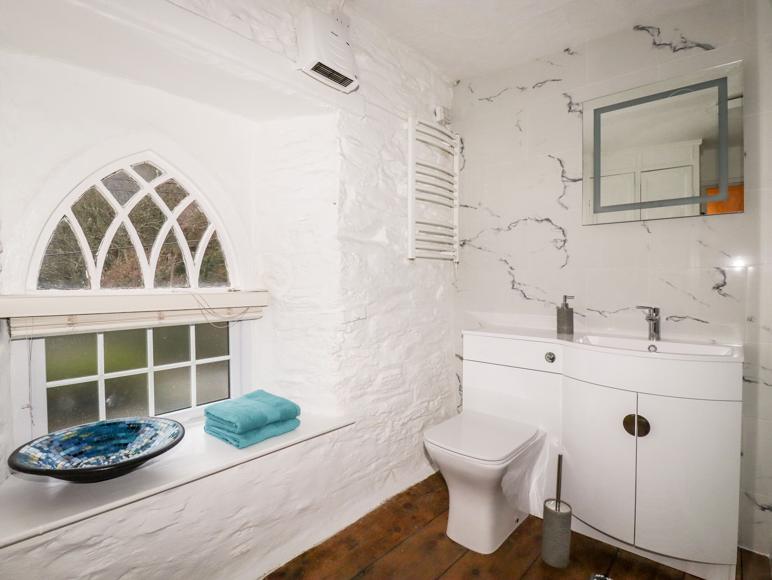 A bathroom with a white marble wall, a white toilet, a white sink, a mirror, and a window with a white frame.