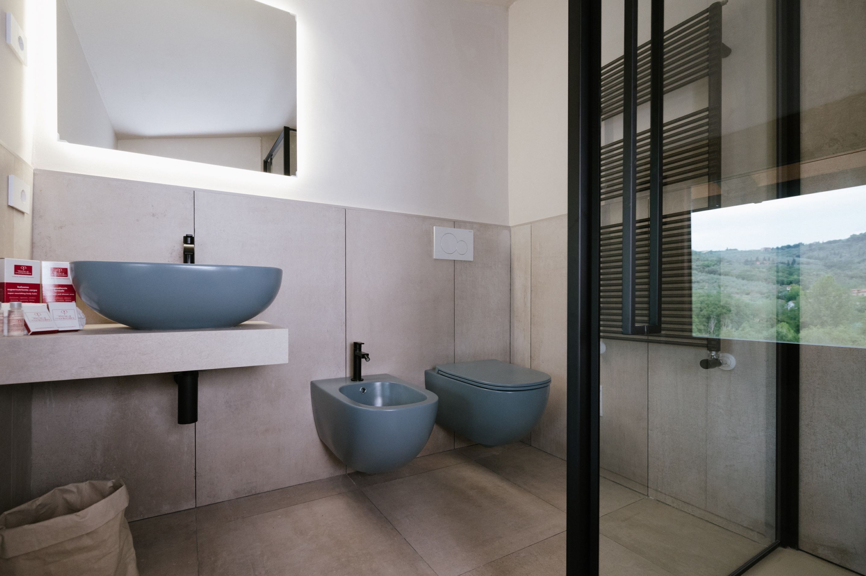 A modern bathroom with a large blue sink, a bidet, and a toilet. There is a mirror above the sink and a window with a view of greenery.