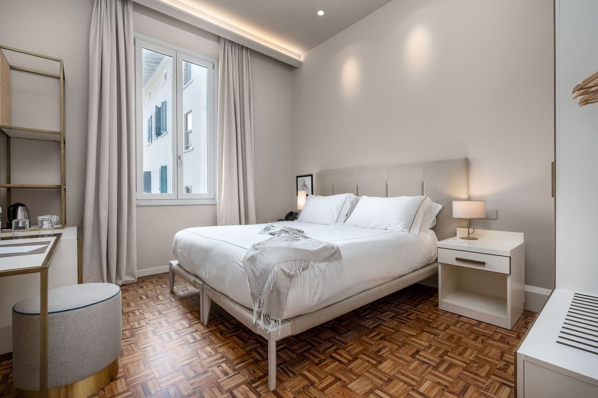 Habitación de hotel moderna con una cama grande, mesitas de noche, un escritorio y vistas a edificios a través del ventanal.