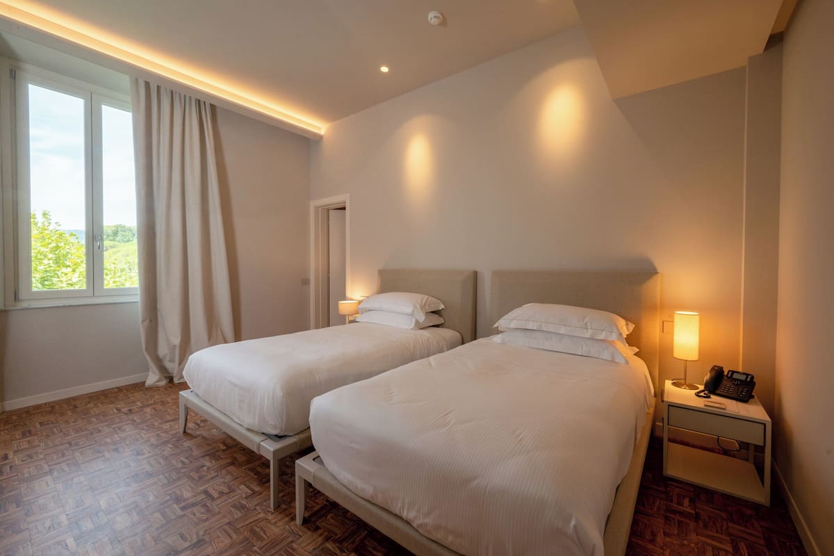 Habitación de hotel con dos camas, una mesita de noche con lámpara y teléfono, una ventana con cortinas y piso de madera.