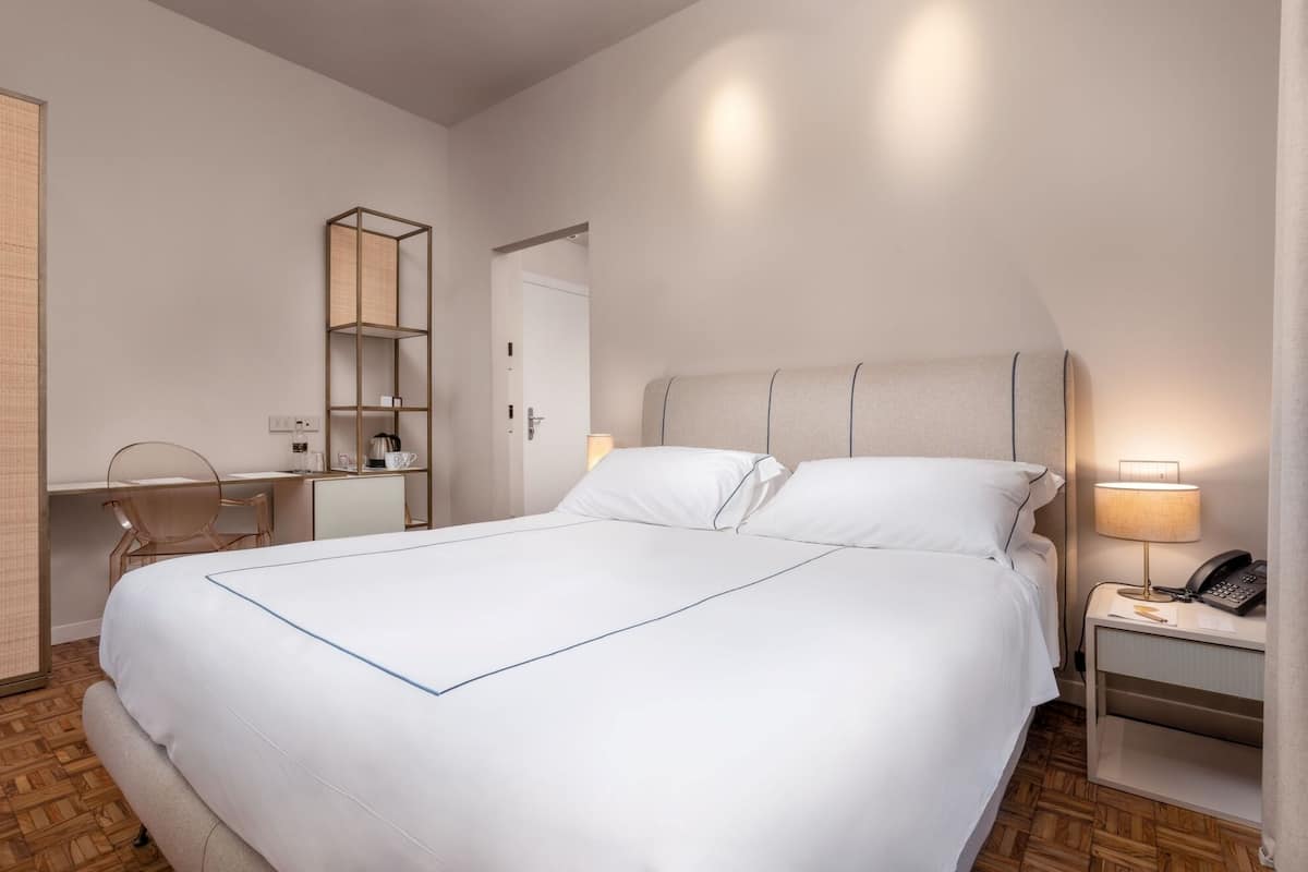 Habitación de hotel con una cama grande, un escritorio con silla, una mesita de noche con lámpara y un armario.