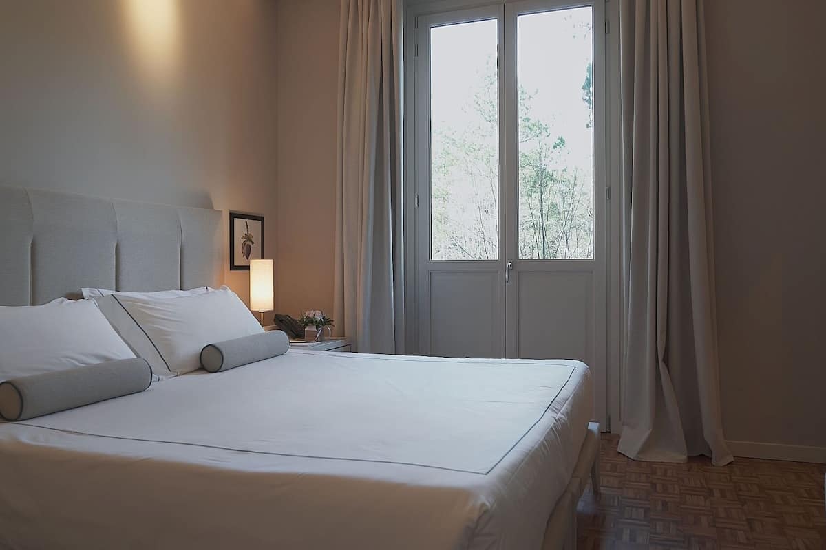 Una cama bien tendida en una habitación de hotel, con un ventanal amplio, una mesita de noche con lámpara y vista a árboles.