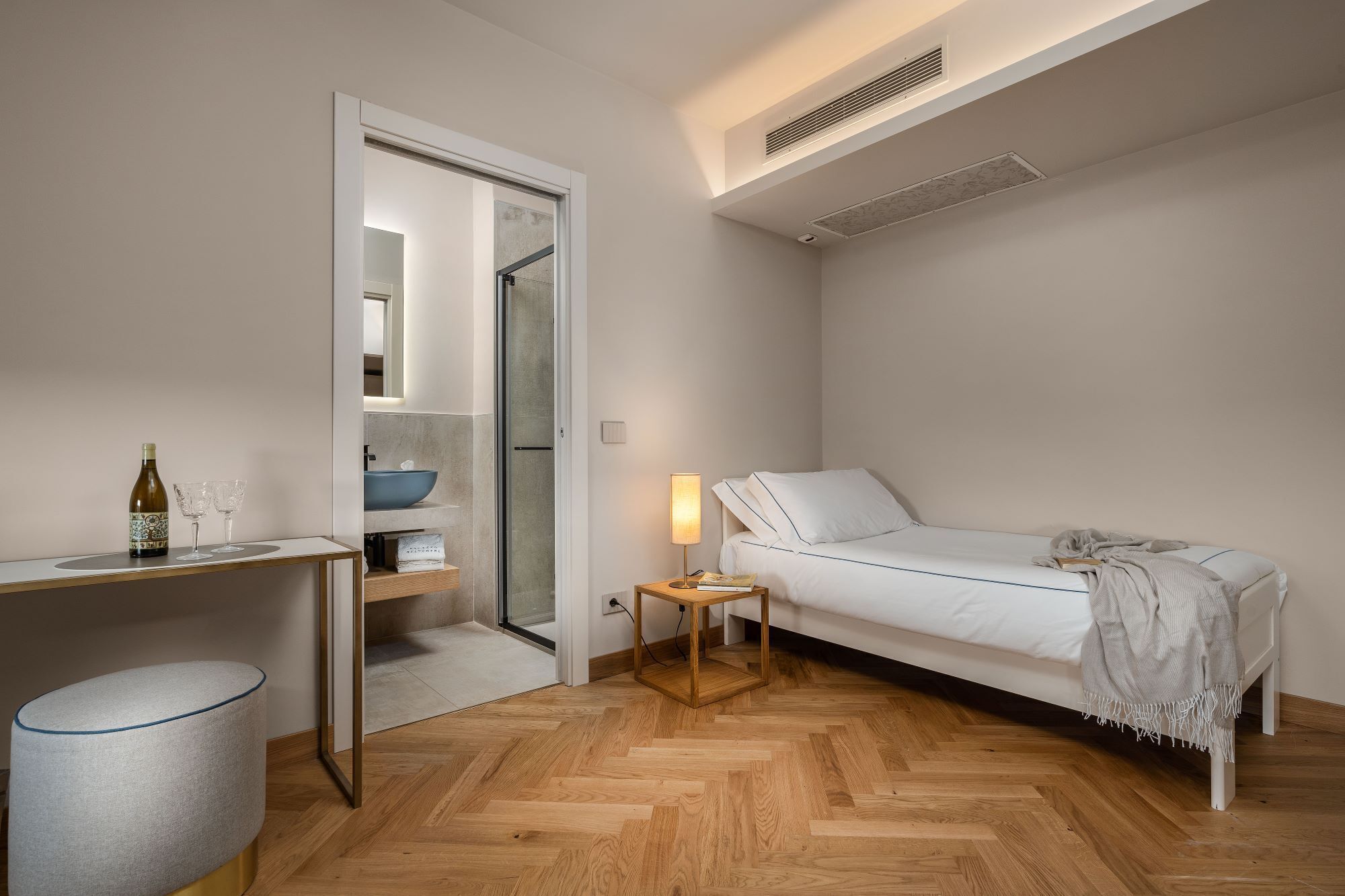 Une chambre moderne avec un lit blanc, une table de chevet avec une lampe, un plancher en bois et une salle de bain avec un lavabo et un miroir.