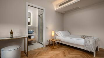 Une chambre moderne avec un lit blanc, une table de chevet avec une lampe, un plancher en bois et une salle de bain avec un lavabo et un miroir.