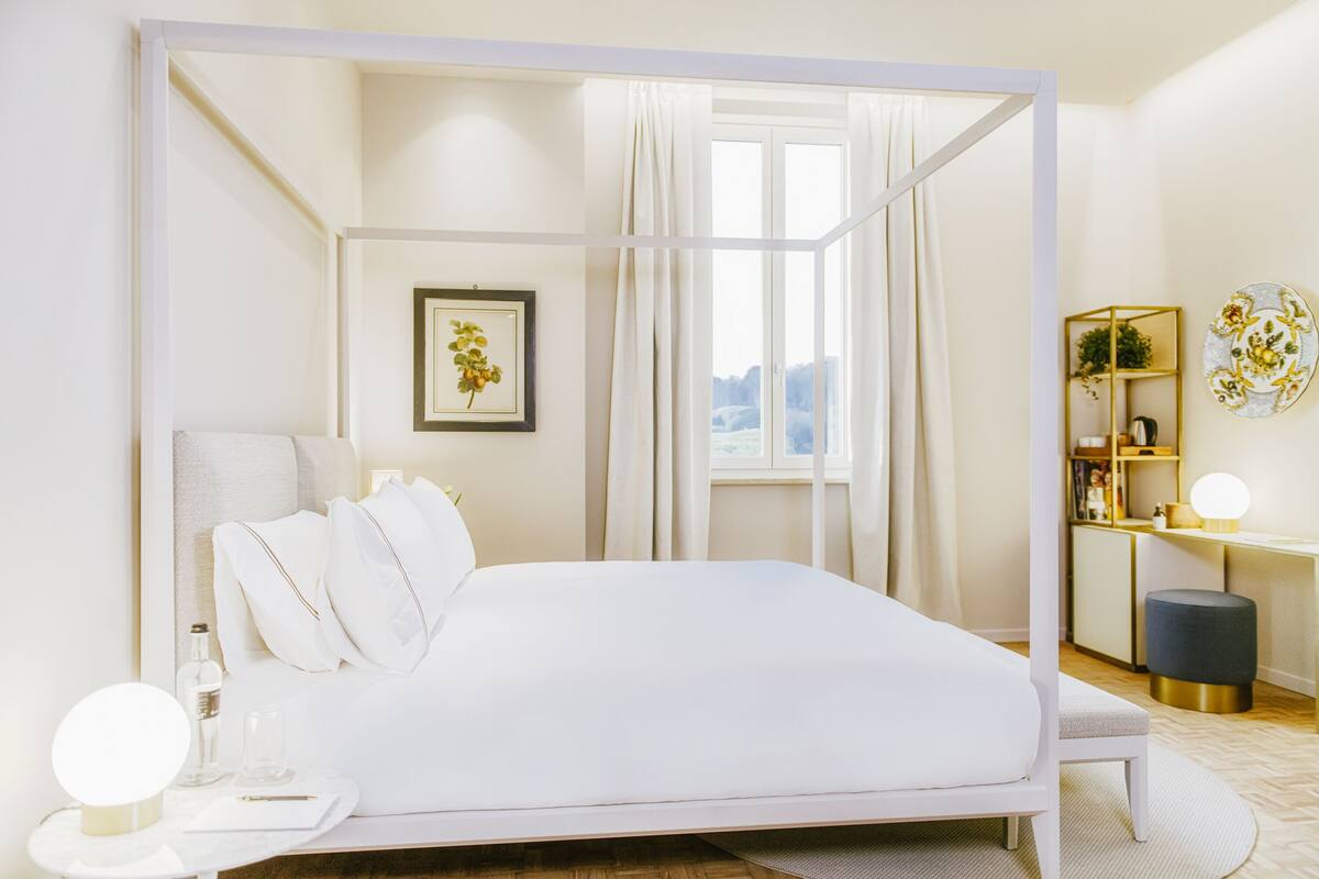 Un dormitorio moderno con una cama grande, una mesita de noche con lámpara, un pequeño escritorio con silla y una ventana con cortinas.