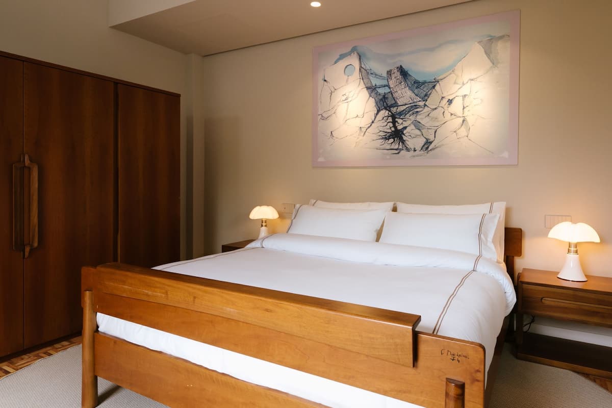 Un dormitorio con cama de madera, dos mesitas de noche con lámparas, un armario de madera grande y un cuadro abstracto colgado en la pared.