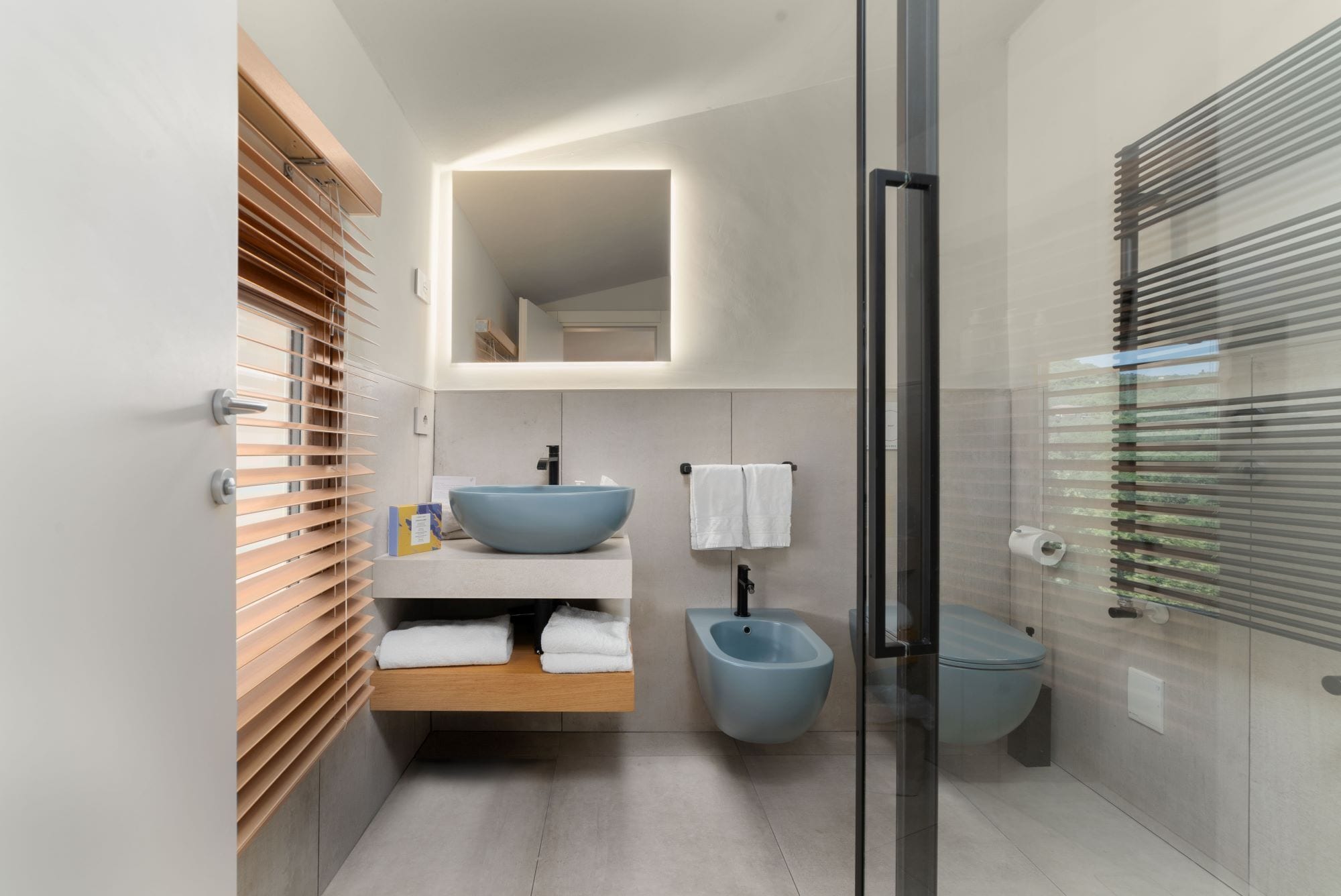 A modern bathroom with a large mirror, a blue sink, a bidet, and a glass shower enclosure.