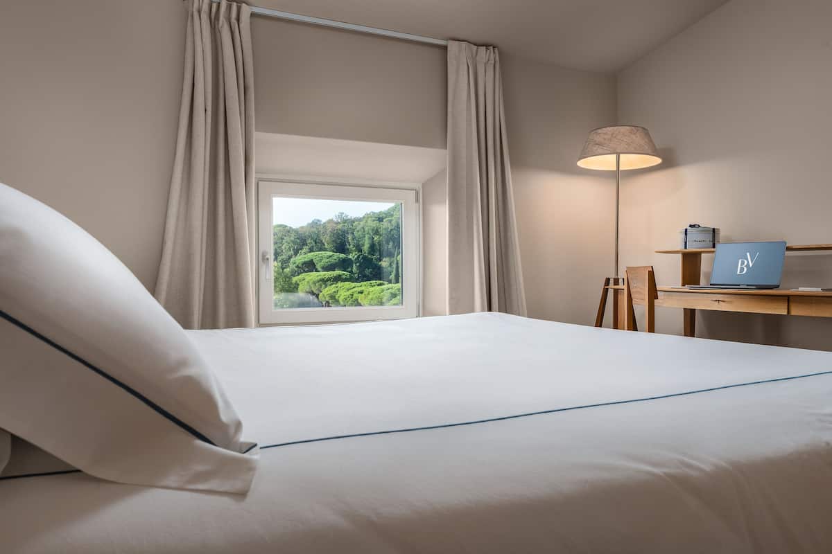Habitación de hotel con cama, escritorio, lámpara y vistas a zonas verdes.