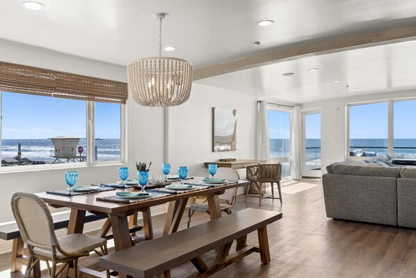 Een ruime eetruimte met uitzicht op de oceaan, een kroonluchter en een moderne woonkamer met een grijze bank.