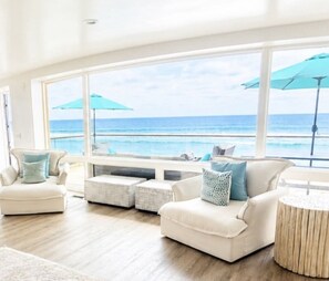A spacious room with large windows overlooking the ocean, furnished with a sofa, ottoman, and a round side table.
