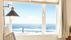 A room with a large window overlooking the sea, a bedside lamp, and a plant on a bedside table.