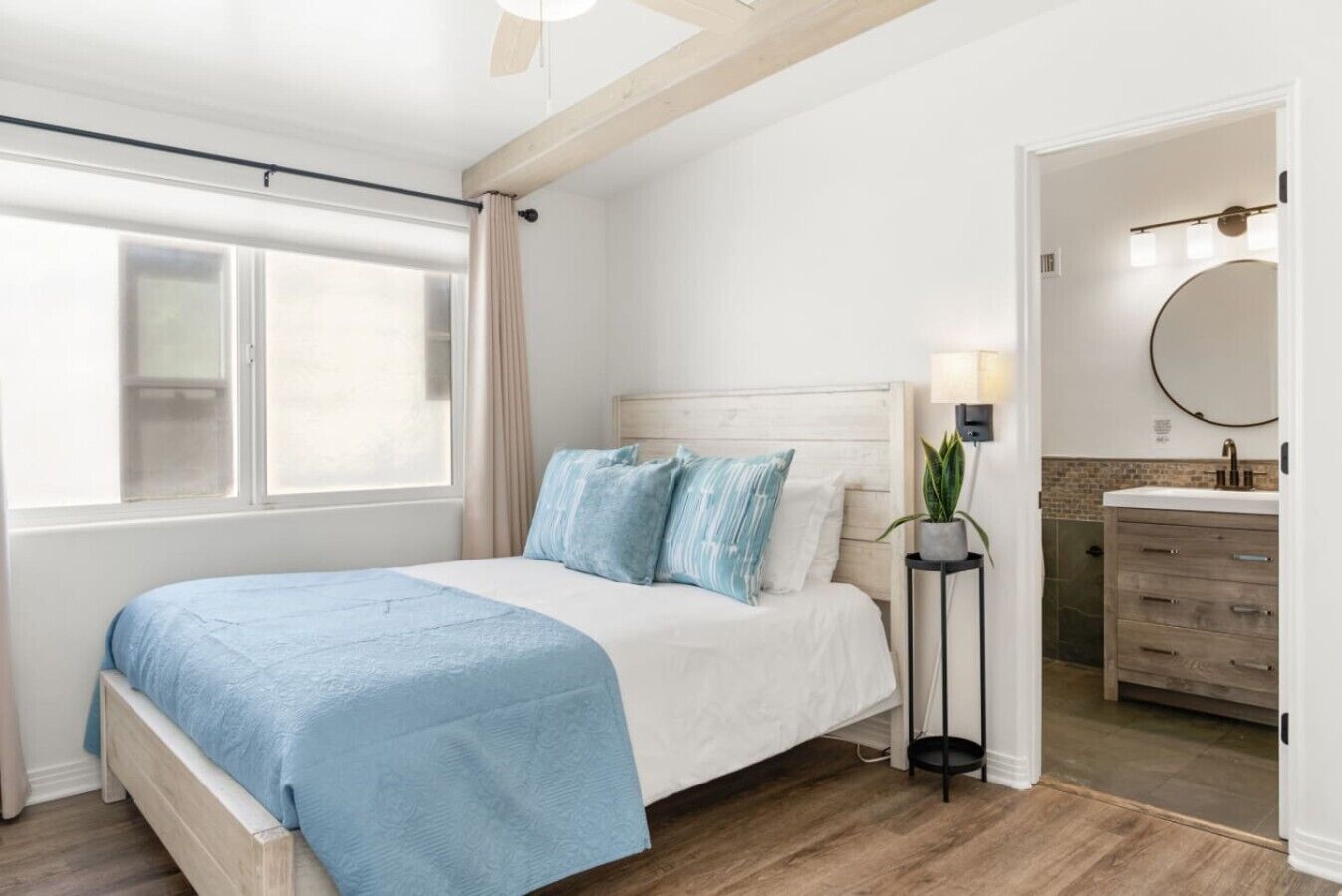 A bedroom with a bed, a bedside table with a lamp, a window with curtains, and a view of a bathroom.