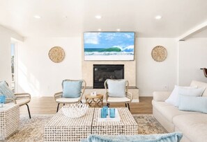 A living room with a fireplace, two armchairs, a sofa, and a television displaying a beach scene.