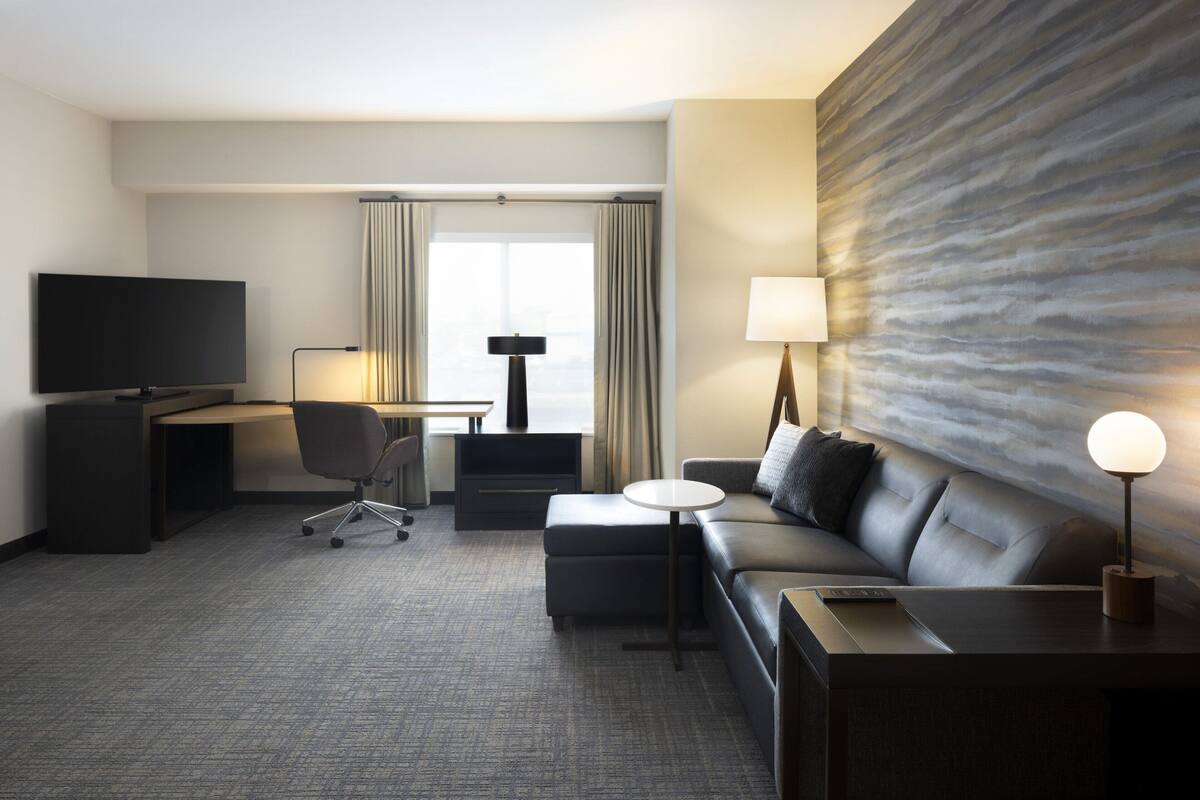 A modern hotel room with a flat-screen TV, a desk with a lamp, a sofa, and a large abstract wall mural.