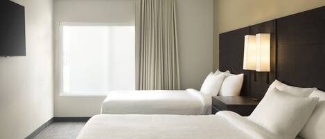 A hotel room with two beds, a TV on the wall, a window with curtains, and a nightstand with a lamp.