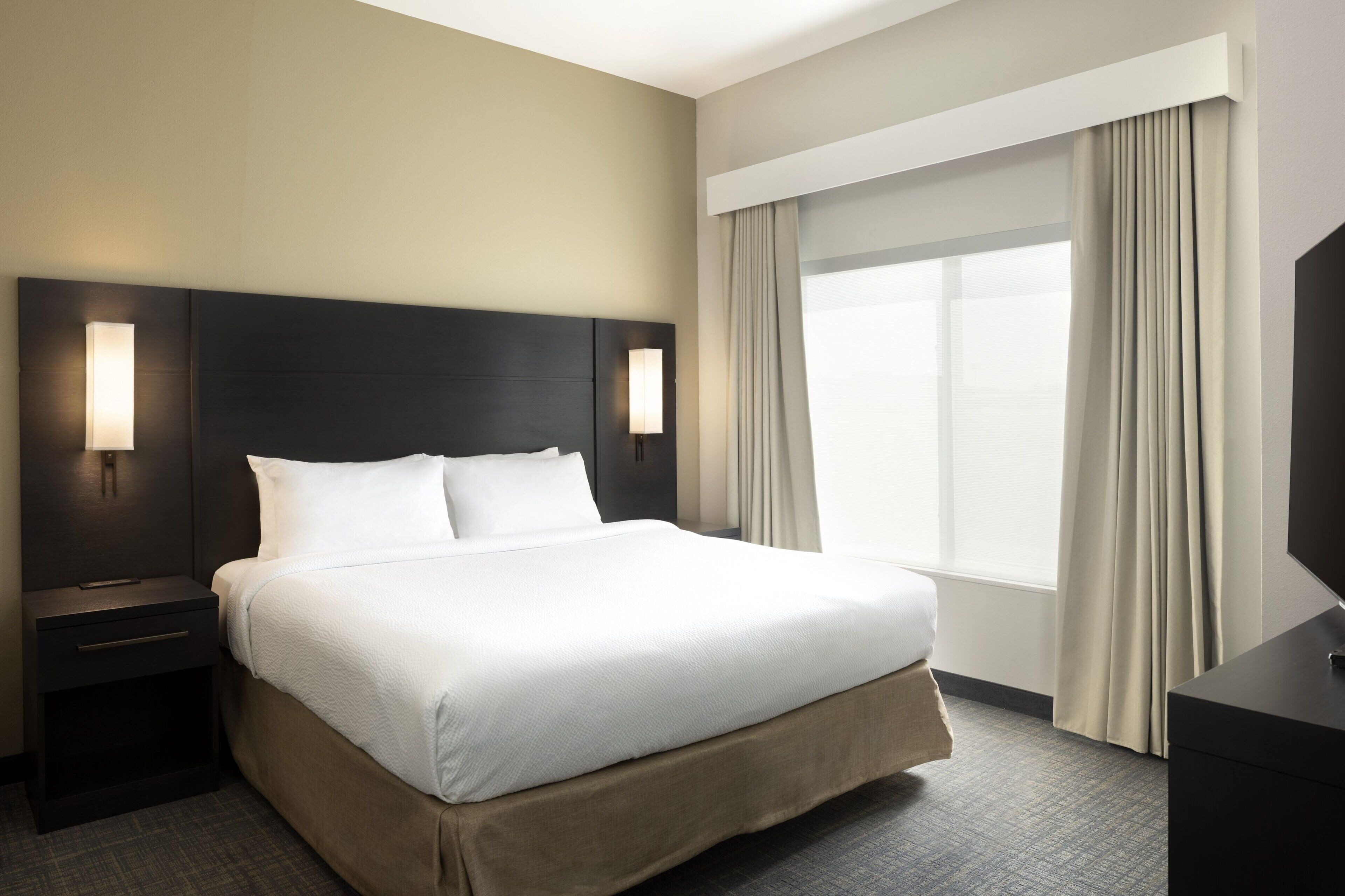 Une chambre d’hôtel avec un grand lit, deux tables de chevet avec des lampes, un téléviseur à écran plat et une grande fenêtre avec des rideaux.