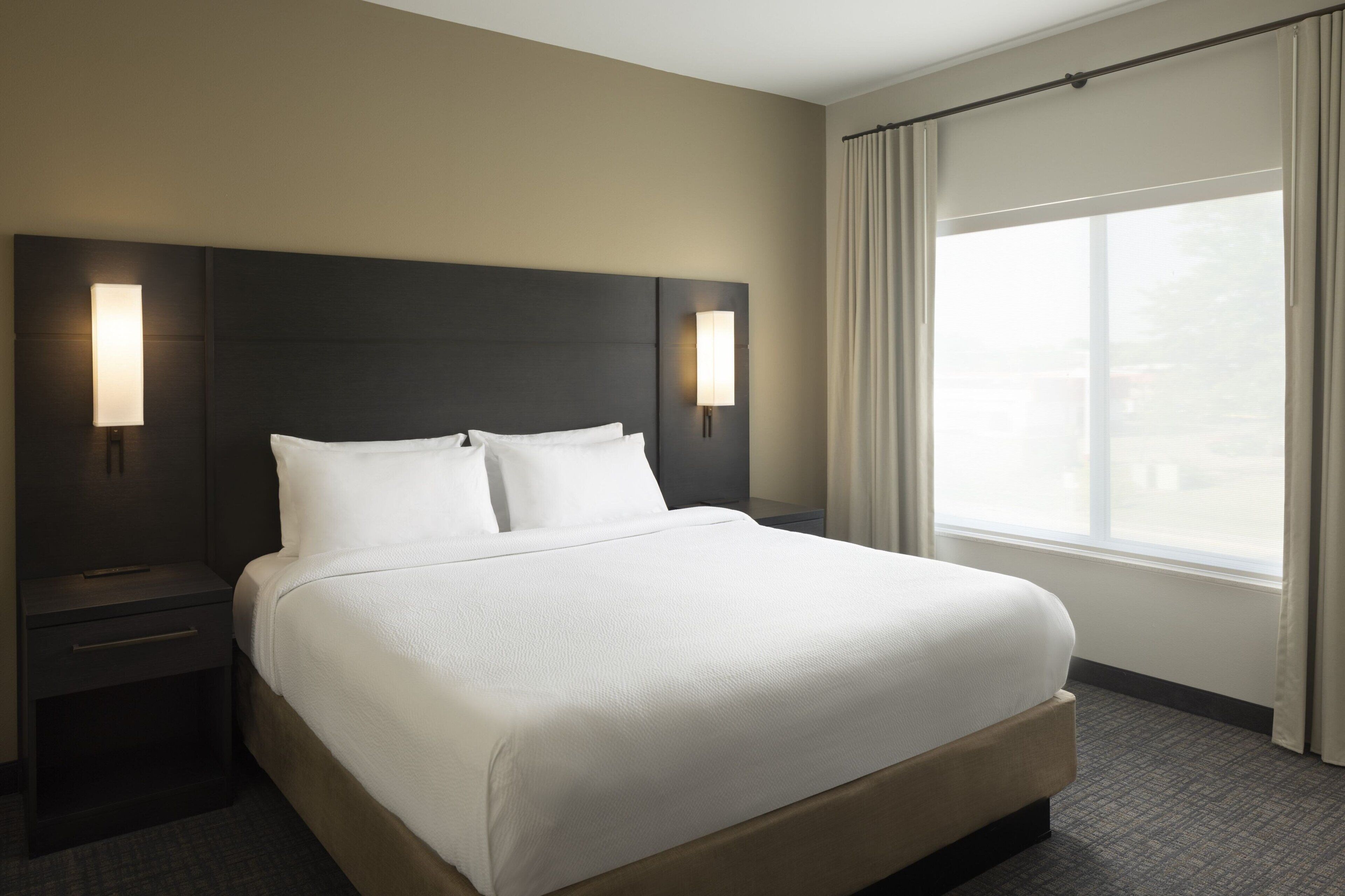 A hotel room with a large bed, two bedside lamps, a window with curtains, and a headboard.