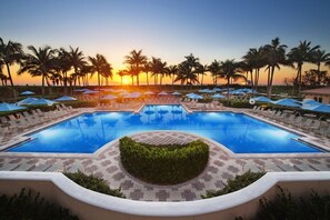 Ein großer Pool mit Liegestühlen und Sonnenschirmen, umgeben von Palmen und Blick auf den Sonnenuntergang.