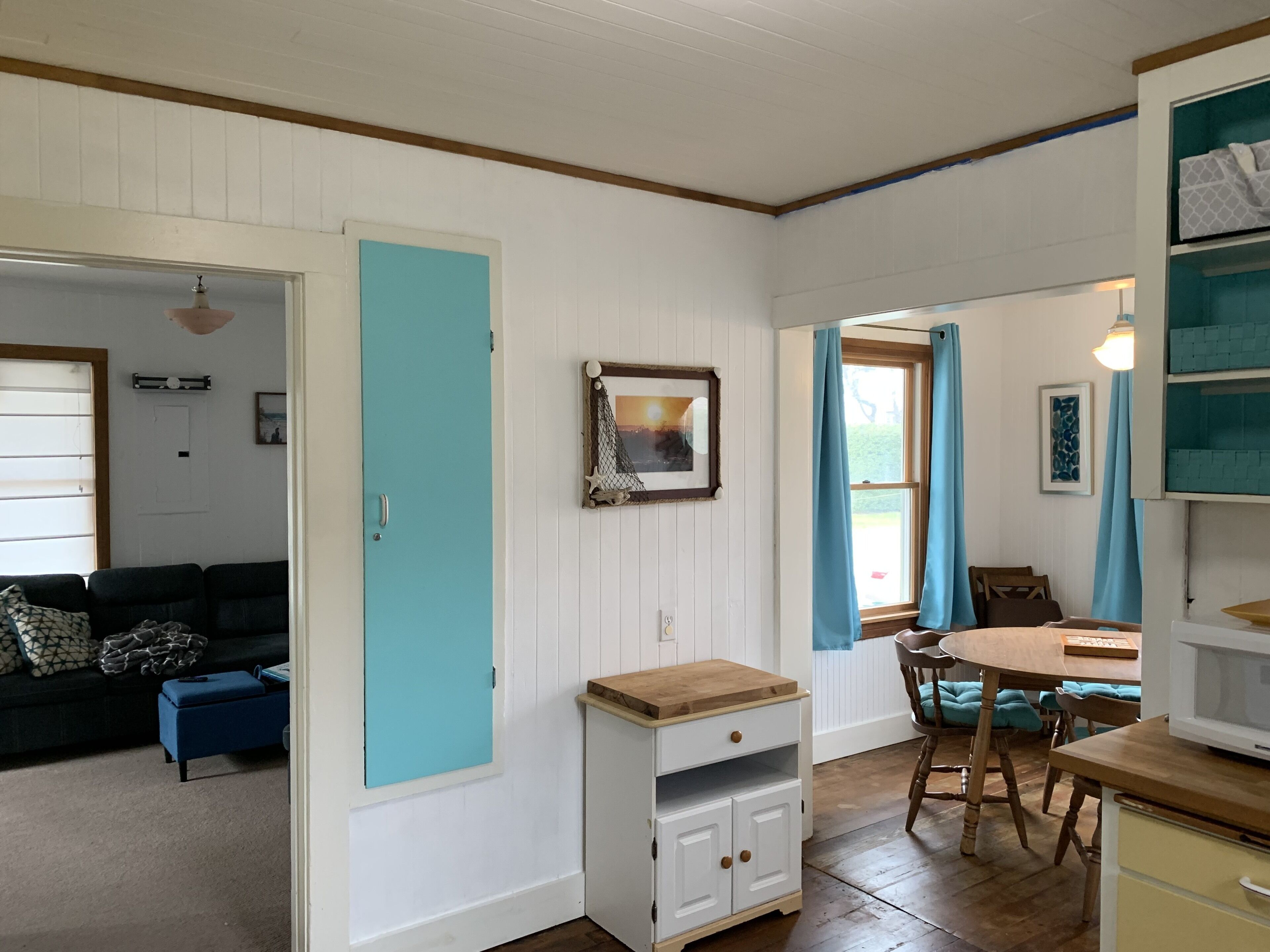 A room with a teal door, a dining table with chairs, a small cabinet, and a window with curtains.