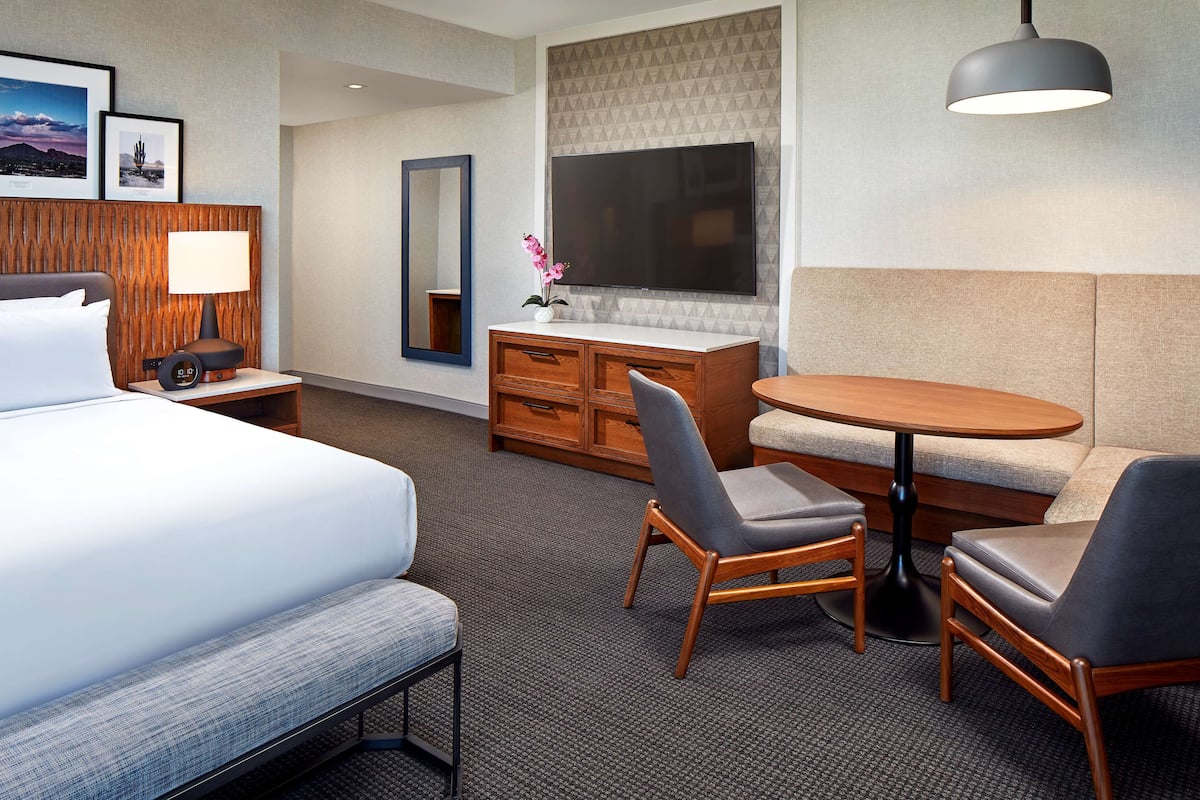 Ein Hotelzimmer mit Bett, Schreibtisch mit Lampe, Kommode, Fernseher, Sofa und rundem Tisch mit zwei Stühlen.