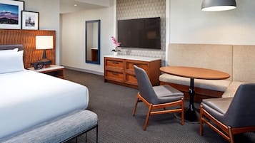 Quarto de hotel com cama, escrivaninha com luminária, cômoda, televisão, sofá e mesa redonda com duas cadeiras.