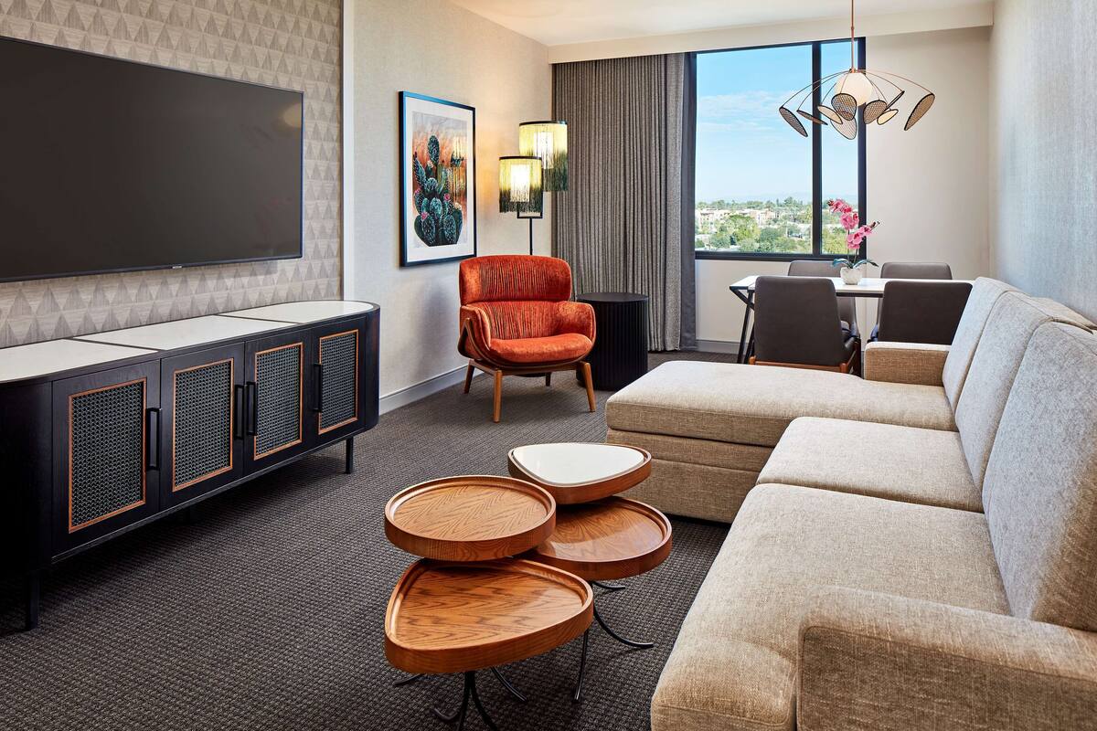 Ein modernes Hotelzimmer mit einem großen Flachbildfernseher, einem roten Samt-Sessel, einem beigen Sofa und einer Essecke mit Tisch und Stühlen.