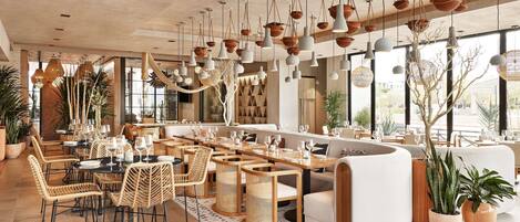 A spacious restaurant with high ceilings, hanging pendant lights, wicker furniture, and large windows offering views of the outdoors.