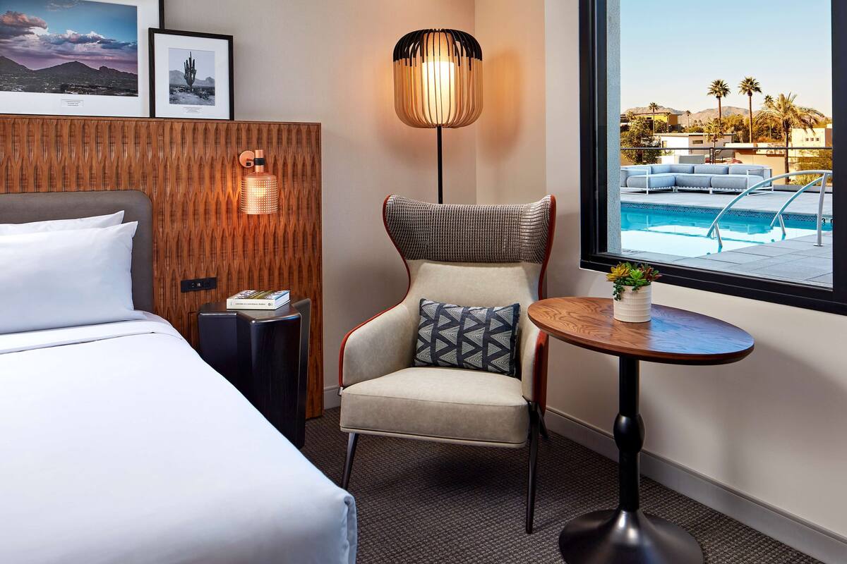Ein Hotelzimmer mit einem Bett, einem Sessel mit Kissen, einem kleinen Tisch mit Pflanze, einer Stehlampe und einem Fenster mit Blick auf einen Pool.