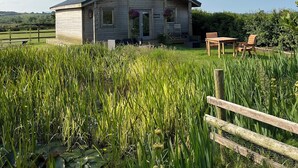 Una cabaña de madera rústica con techo inclinado, rodeada por un campo verde exuberante y una valla de madera.