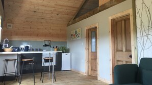 Una cocina con vigas de madera en el techo, encimera blanca y taburetes de bar. Hay una puerta que da a otra habitación y una silla verde al frente.