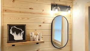 A bathroom with wooden paneling, a framed picture of a tree silhouette, a mirror, and a sink with a faucet. - The BL Cottage - Lake Getaway with Panoramic Views, near Cascade Mountain (Montello)