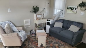 A living room with a beige armchair, a grey sofa, a patterned rug, a small white table, and a wall-mounted heater. - Little House (Monticello)