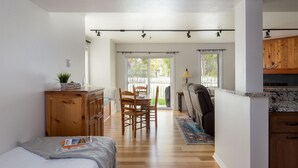 Una habitación amplia con cama, cómoda de madera, zona de comedor con sillas, cocina con armarios de madera y un gran ventanal corredero que da a un espacio exterior.